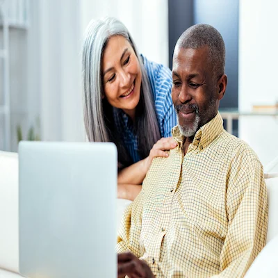 Man smiles while sitting and using a laptop while woman smiles behind him and pats him on the shoulder