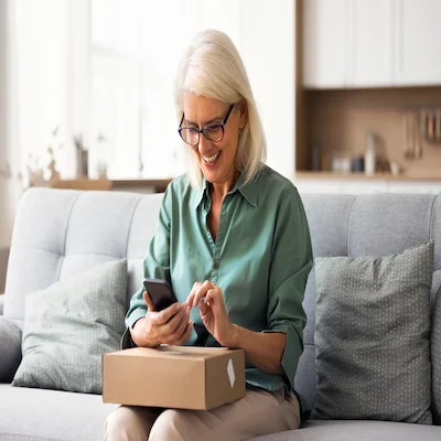 Older woman smiles while looking at her phone and holding a newly arrived package in her lap