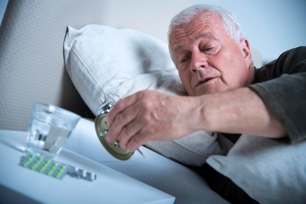 Senior man in bed in the morning. He is looking at alarm clock to see what is the time. On a cabinet there are some pills and glass of water.
