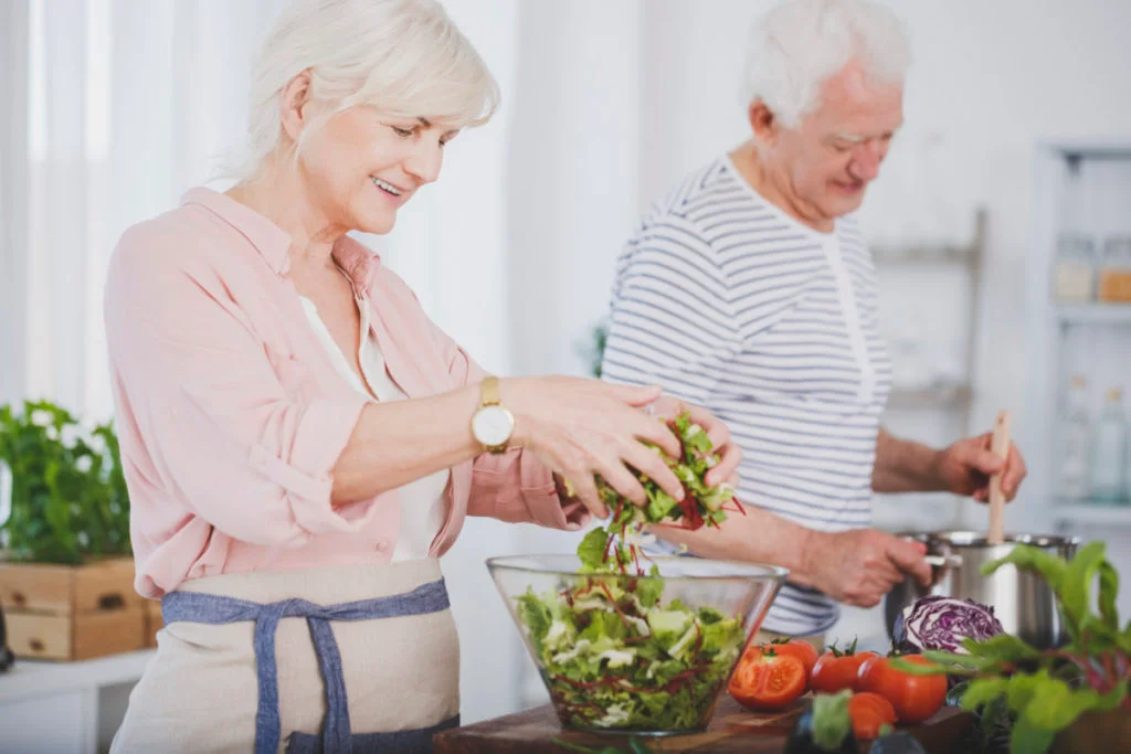 Older woman tossing a salad and her husband stirring food in a pot, cooking together 
